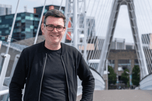 Man smiling outdoors with cityscape and modern bridge in background.