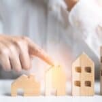 Person pointing at increasing wooden house models for property growth.